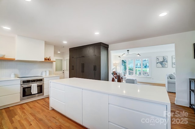 kitchen with black electric stovetop, white cabinets, a kitchen island, stainless steel oven, and light wood-type flooring