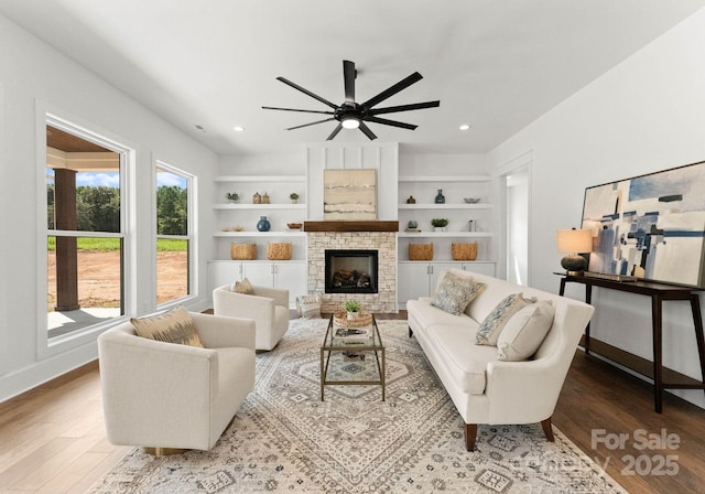 living room featuring built in shelves, light hardwood / wood-style flooring, a stone fireplace, and ceiling fan