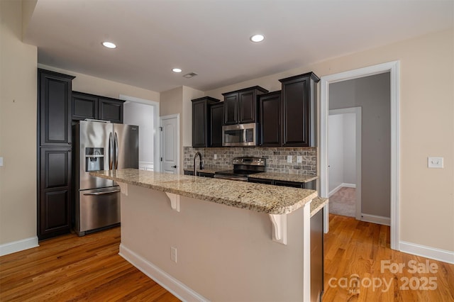 kitchen with a breakfast bar, appliances with stainless steel finishes, an island with sink, light stone countertops, and light hardwood / wood-style floors