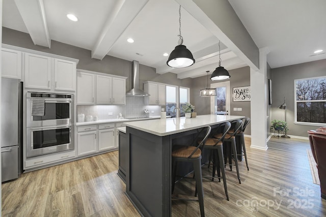 kitchen with a center island with sink, appliances with stainless steel finishes, white cabinetry, pendant lighting, and wall chimney exhaust hood