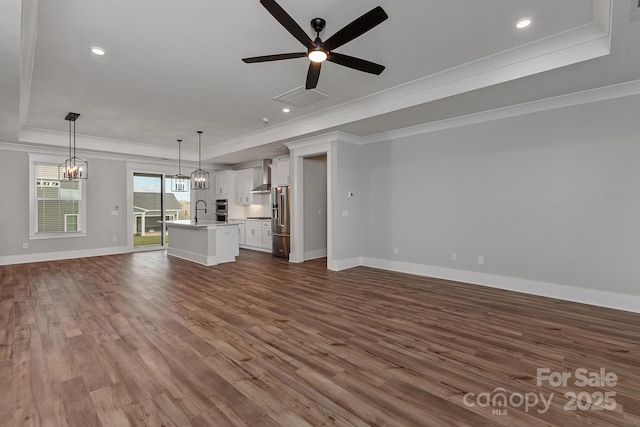 unfurnished living room featuring sink, crown molding, a tray ceiling, hardwood / wood-style flooring, and ceiling fan with notable chandelier