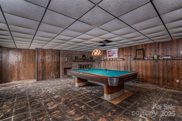 recreation room featuring a paneled ceiling, wooden walls, and a brick fireplace