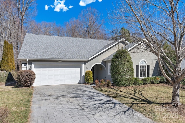 ranch-style home with an attached garage, a shingled roof, stucco siding, a front lawn, and decorative driveway