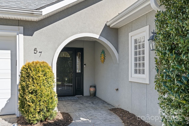 view of exterior entry with stucco siding, an attached garage, and a shingled roof