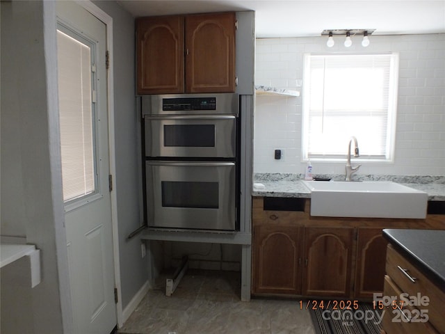 kitchen with tasteful backsplash, stainless steel double oven, sink, and light stone counters