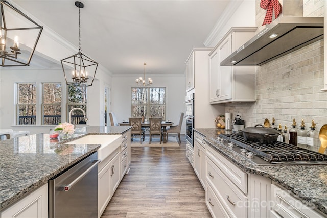 kitchen featuring stainless steel appliances, white cabinetry, wall chimney range hood, dark stone countertops, and pendant lighting