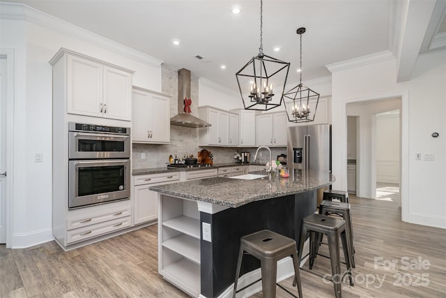 kitchen with stainless steel appliances, white cabinets, wall chimney range hood, open shelves, and a center island with sink