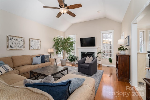 living area featuring baseboards, vaulted ceiling, a premium fireplace, light wood-style flooring, and a ceiling fan