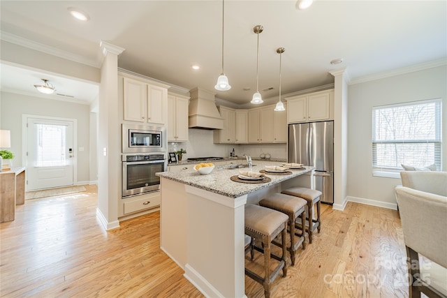 kitchen with a breakfast bar area, custom exhaust hood, light stone counters, appliances with stainless steel finishes, and a kitchen island with sink