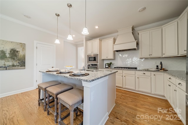 kitchen with light stone counters, hanging light fixtures, an island with sink, custom range hood, and stainless steel appliances