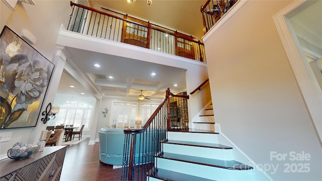 stairs with coffered ceiling, ceiling fan with notable chandelier, a towering ceiling, beamed ceiling, and wood finished floors