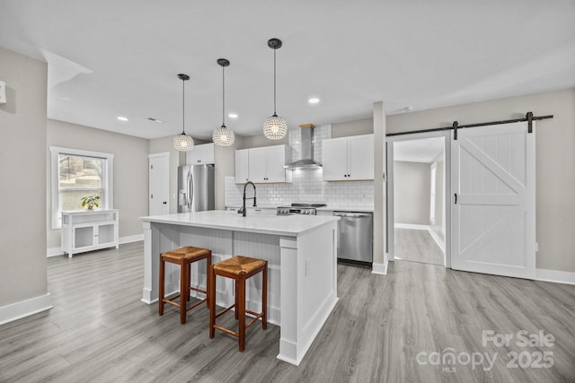 kitchen with hanging light fixtures, appliances with stainless steel finishes, an island with sink, wall chimney range hood, and white cabinets