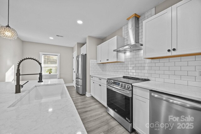 kitchen featuring wall chimney range hood, sink, stainless steel appliances, white cabinets, and decorative light fixtures