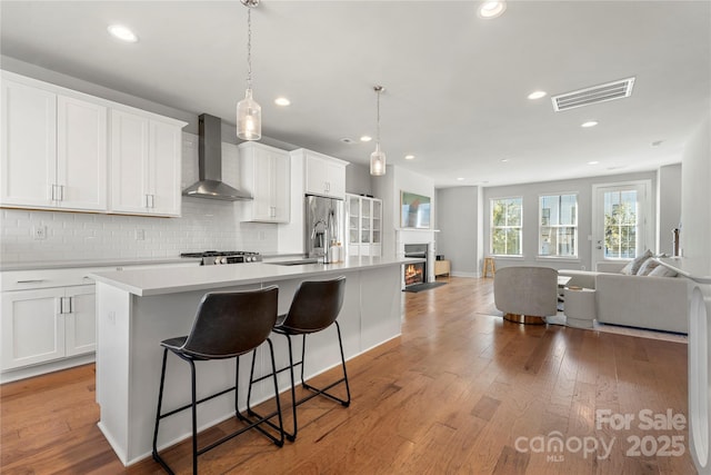 kitchen with wall chimney range hood, light hardwood / wood-style flooring, a kitchen island with sink, white cabinets, and decorative backsplash