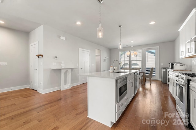 kitchen with sink, a kitchen island with sink, stainless steel appliances, white cabinets, and decorative light fixtures