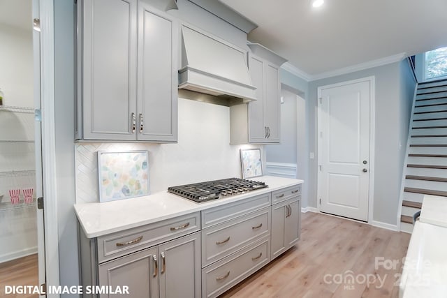 kitchen with stainless steel gas cooktop, custom exhaust hood, gray cabinetry, crown molding, and light hardwood / wood-style floors