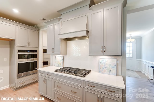 kitchen with gray cabinetry, crown molding, custom range hood, stainless steel appliances, and light hardwood / wood-style floors