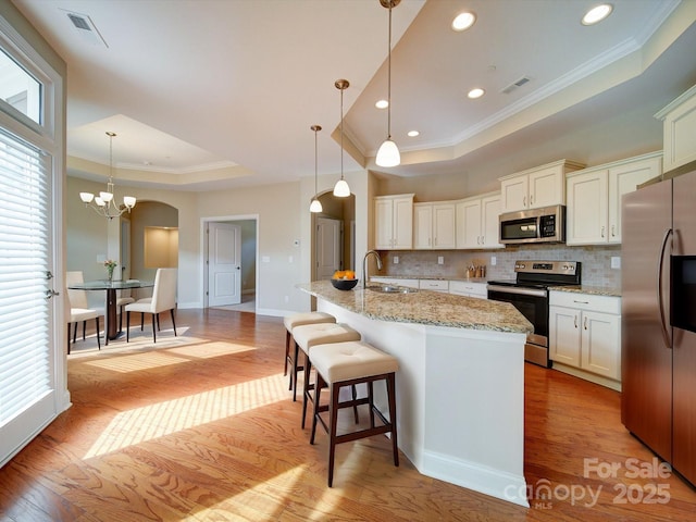 kitchen featuring stainless steel appliances, light stone countertops, a center island with sink, decorative light fixtures, and a raised ceiling