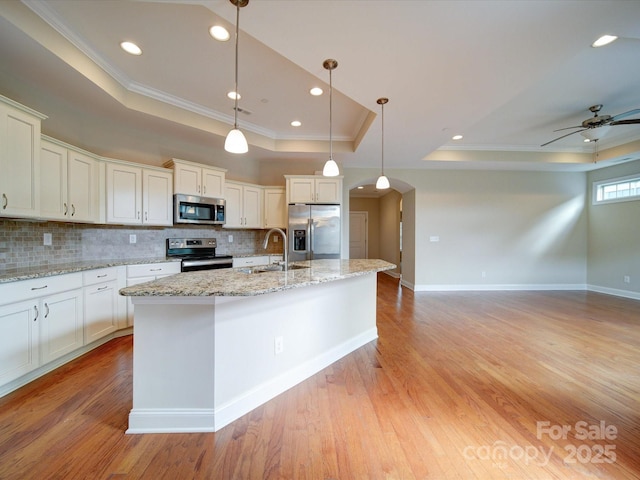 kitchen with stainless steel appliances, a raised ceiling, hanging light fixtures, and a center island with sink