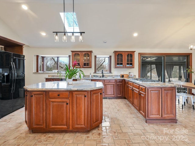 kitchen with hanging light fixtures, a center island, light stone counters, stainless steel gas cooktop, and black fridge with ice dispenser