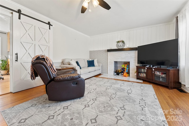 living room with a brick fireplace, wood-type flooring, ceiling fan, a barn door, and ornamental molding