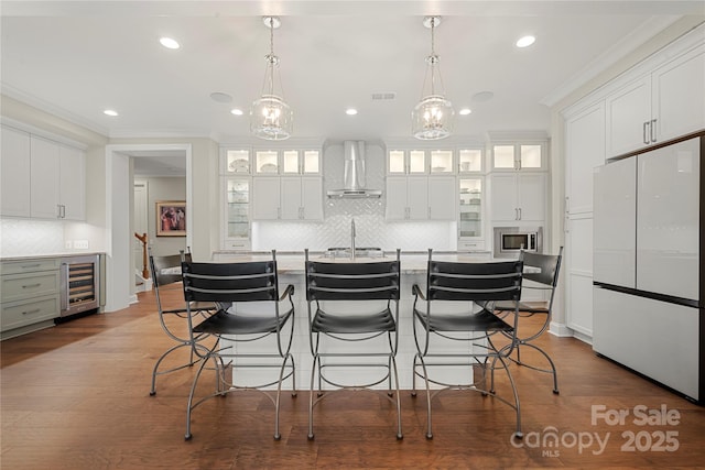 kitchen featuring stainless steel microwave, white cabinetry, white fridge, a kitchen island with sink, and wall chimney exhaust hood