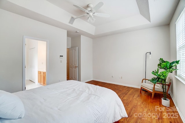 bedroom with a raised ceiling, light wood-type flooring, ensuite bathroom, and ceiling fan