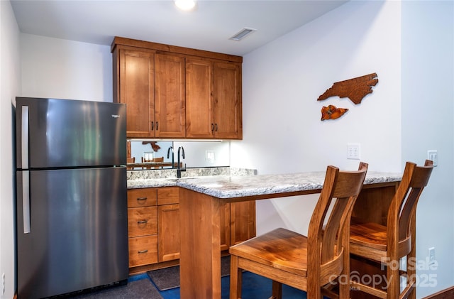 kitchen with a kitchen bar, sink, light stone counters, stainless steel refrigerator, and kitchen peninsula