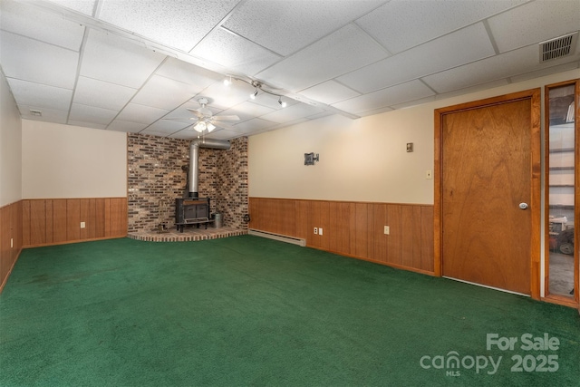 basement with dark colored carpet, a wood stove, a drop ceiling, and baseboard heating