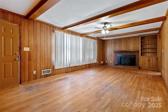 unfurnished living room featuring ceiling fan, visible vents, wooden walls, a wood stove, and beam ceiling