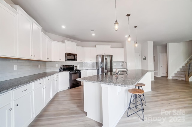 kitchen with white cabinetry, decorative light fixtures, an island with sink, dark stone counters, and black appliances