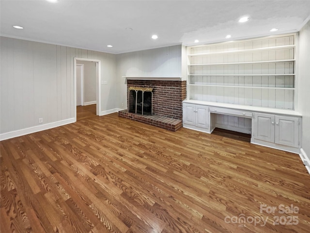 unfurnished living room with built in desk, wood walls, wood-type flooring, crown molding, and a brick fireplace