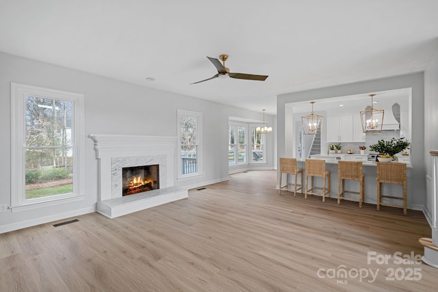 unfurnished living room with a high end fireplace, ceiling fan with notable chandelier, and light wood-type flooring