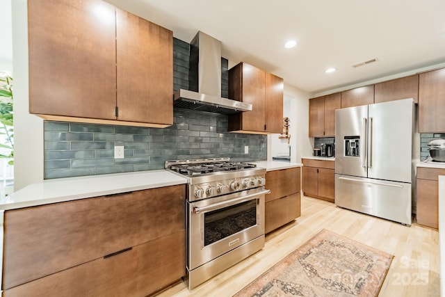 kitchen featuring stainless steel appliances, wall chimney range hood, light wood-type flooring, and light countertops