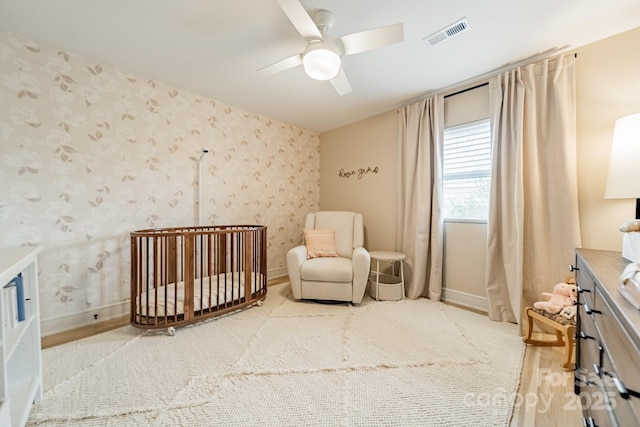 bedroom with visible vents, ceiling fan, a crib, baseboards, and wallpapered walls