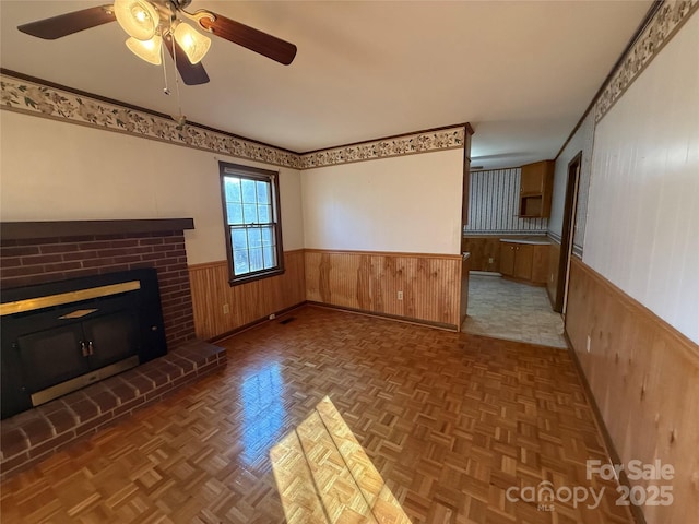 unfurnished living room featuring ceiling fan, a brick fireplace, dark parquet floors, and wood walls