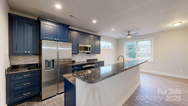 kitchen featuring tasteful backsplash, appliances with stainless steel finishes, a center island with sink, and dark stone counters