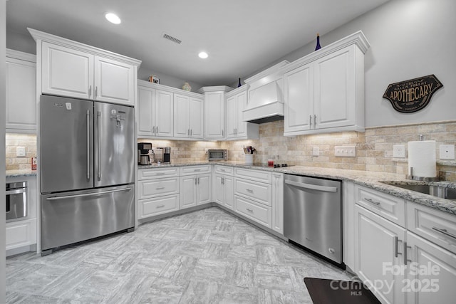 kitchen featuring white cabinetry, decorative backsplash, stainless steel appliances, light stone countertops, and custom range hood