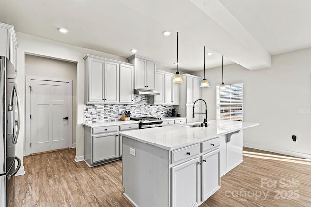 kitchen with decorative light fixtures, an island with sink, sink, light stone counters, and stainless steel appliances