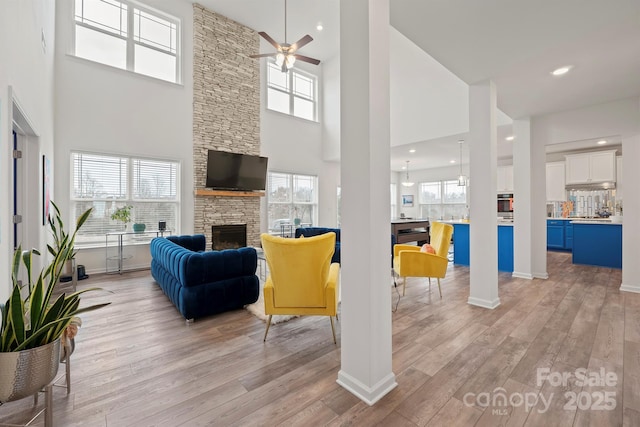 living room with a stone fireplace, light hardwood / wood-style flooring, ceiling fan, and a high ceiling