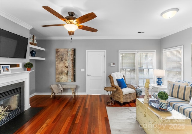 living area with crown molding, a glass covered fireplace, ceiling fan, wood finished floors, and baseboards