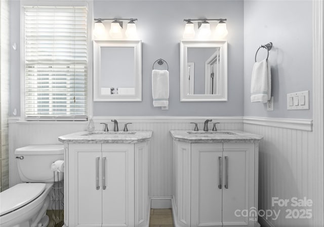 full bath with toilet, a wainscoted wall, two vanities, and a sink