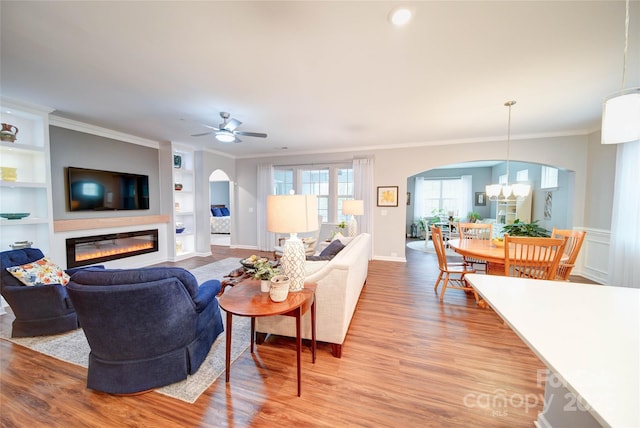 living room featuring built in shelves, light hardwood / wood-style flooring, ornamental molding, and ceiling fan with notable chandelier