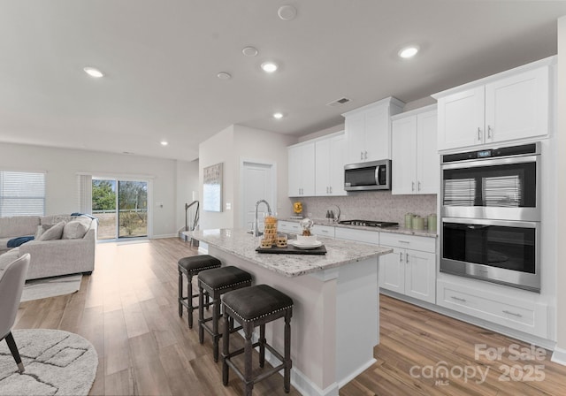 kitchen with a breakfast bar, an island with sink, white cabinetry, stainless steel appliances, and light stone countertops