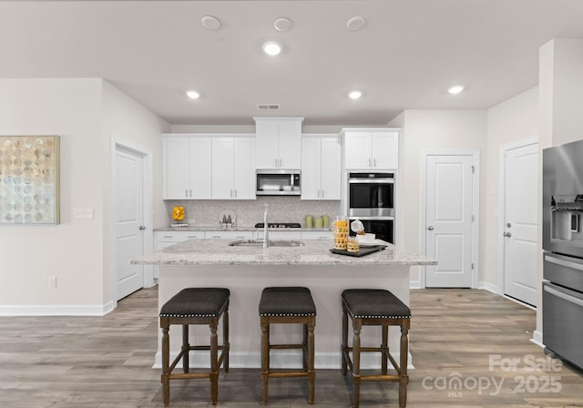 kitchen with sink, appliances with stainless steel finishes, a kitchen island with sink, light stone counters, and white cabinets