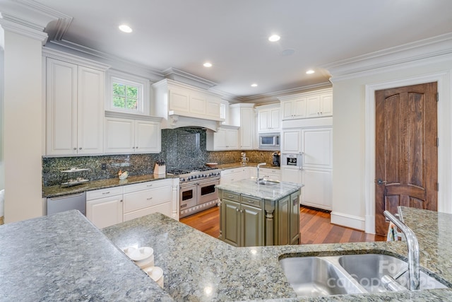 kitchen featuring white cabinets, ornamental molding, a kitchen island with sink, built in appliances, and light stone counters