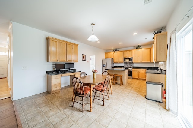 kitchen with light brown cabinetry, decorative light fixtures, built in desk, and stainless steel appliances