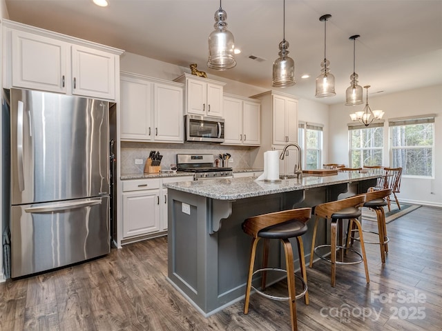 kitchen featuring a kitchen island with sink, white cabinetry, stainless steel appliances, light stone countertops, and decorative light fixtures