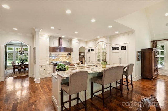 kitchen featuring wall chimney range hood, light stone countertops, a healthy amount of sunlight, a kitchen bar, and a large island with sink