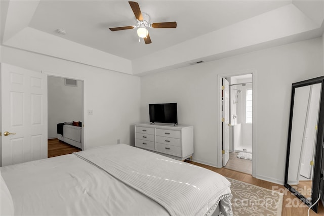 bedroom with ceiling fan, ensuite bath, a tray ceiling, and hardwood / wood-style floors